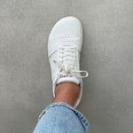 Load image into Gallery viewer, A white low-top sneaker worn on a person's foot, with light blue jeans visible at the cuff, photographed from above against a gray textured floor. The tongue shows the brand name ICARUS.

